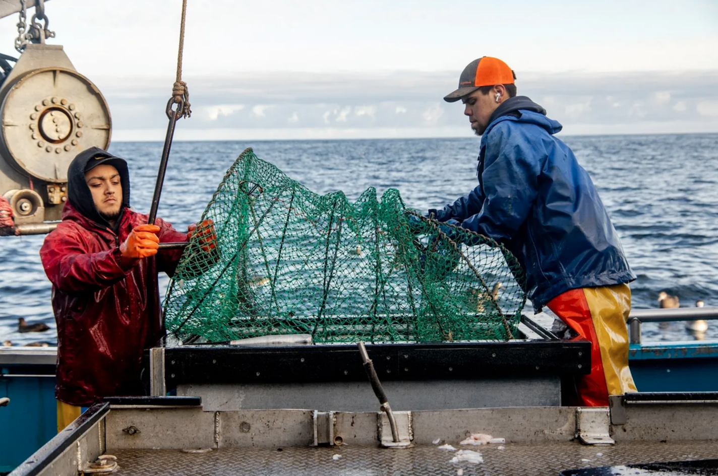 Two men are standing on a fishing boat wearing brightly colored, warm fishing apparel and caps. They are holding a hoop net used for harvesting fish.